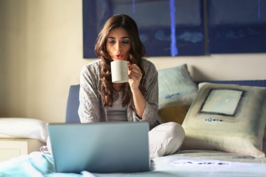 Woman in grey jacket relaxing and sitting on bed uses grey laptop and drinks a hot drink. Photo by Andrea Piacquadio: https://www.pexels.com/photo/woman-in-grey-jacket-sits-on-bed-uses-grey-laptop-935743/